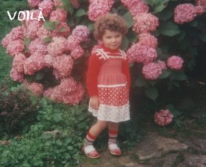A little girl with curly light brown hair, a red and white Summer dress in front of a big pink hortensia
